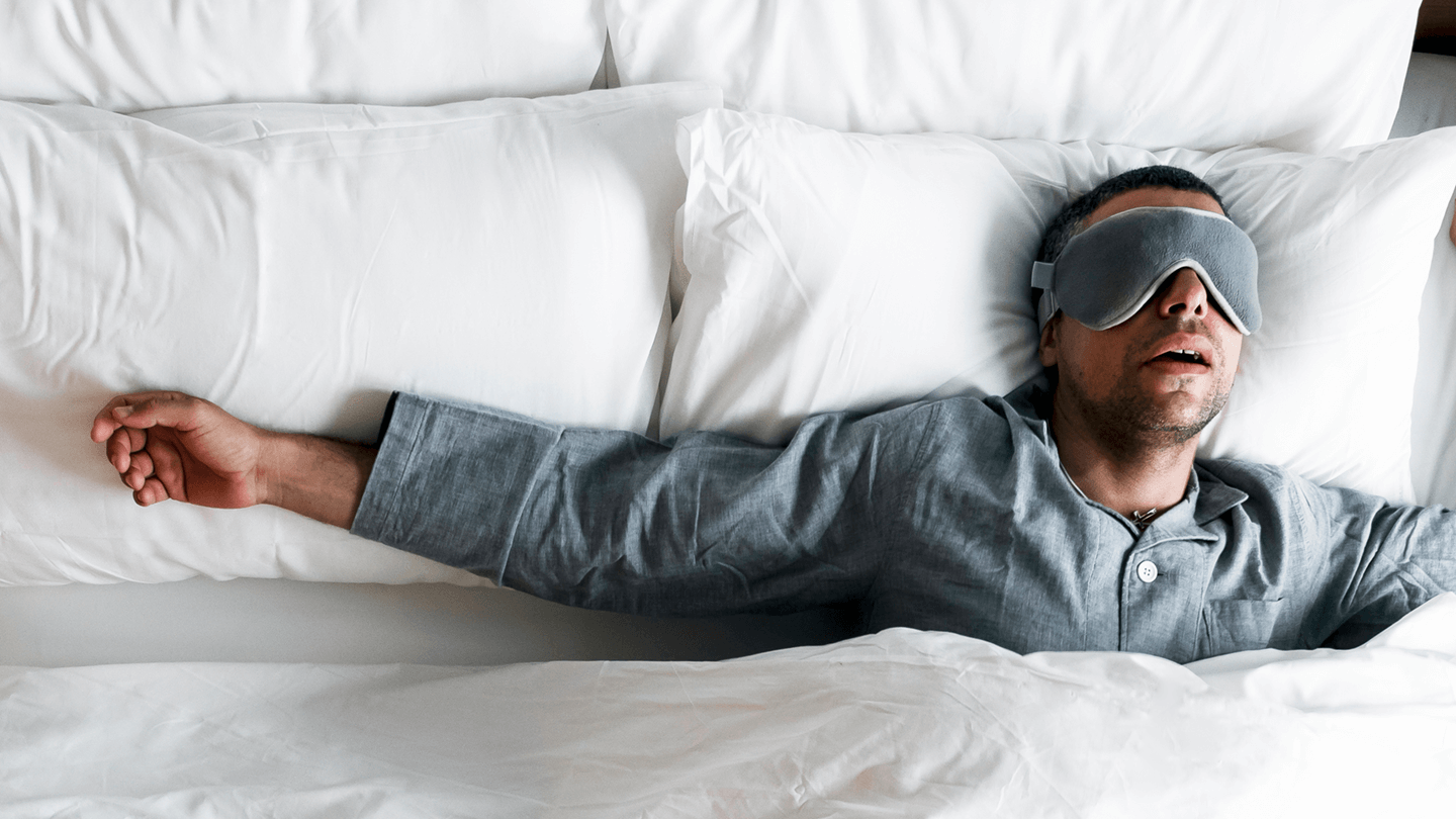 a man with eye mask sleeping on bed and snoring with open mouth